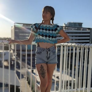 May include: A woman wearing a blue and white crochet crop top with short sleeves and a blue denim skirt. She is standing on a balcony with a white railing and has a black tattoo on her leg.