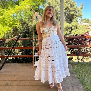 Könnte beinhalten: Ein weißes Häkelkleid mit floral gemustertem Oberteil. Das Kleid hat ein eng anliegendes Oberteil und einen fließenden, durchbrochenen Rock. Die Person trägt weiße Sandalen. Der Hintergrund umfasst eine Holzterrasse, Grün und einen blauen Himmel.