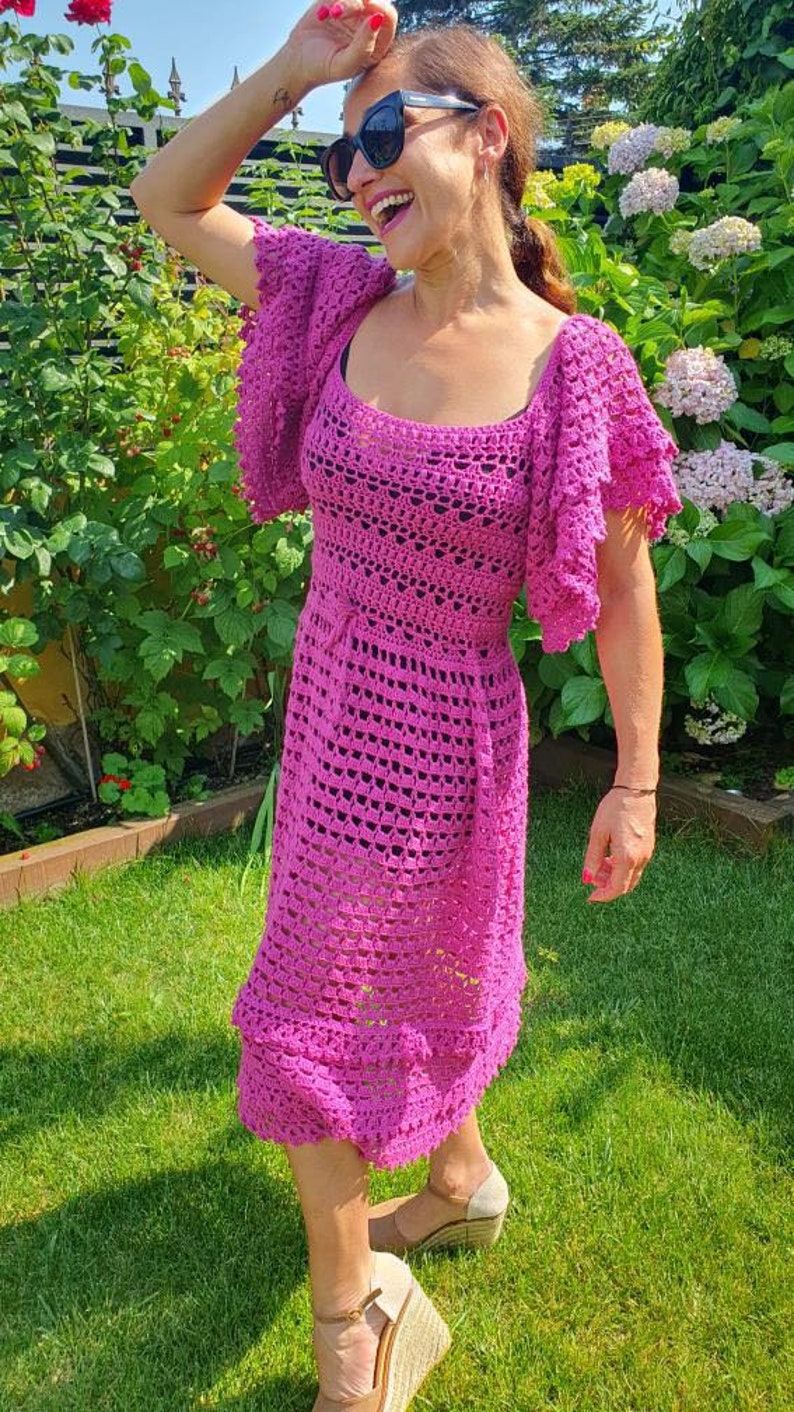 May include: A pink crochet dress with a square neckline and short, ruffled sleeves. The dress is fitted at the waist and flows out to a midi length.