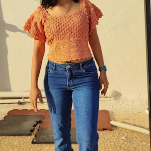 May include: A woman wearing a crocheted orange top with a ruffled neckline and blue jeans with slits at the bottom.