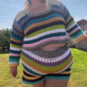 K&ouml;nnte beinhalten: Ein farbenfrohes, gestreiftes H&auml;kel-Zweiteiler-Set. Das lang&auml;rmelige Crop-Top und die passenden Shorts zeigen horizontale Streifen in verschiedenen Farben, darunter Blau, Gr&uuml;n, Gelb, Rosa und Braun. Die handgefertigte H&auml;kelarbeit ist deutlich sichtbar.