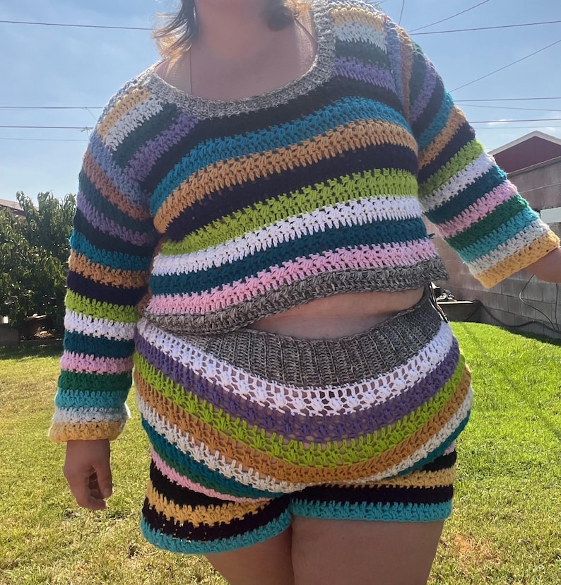 K&ouml;nnte beinhalten: Ein farbenfrohes, gestreiftes H&auml;kel-Zweiteiler-Set. Das lang&auml;rmelige Crop-Top und die passenden Shorts zeigen horizontale Streifen in verschiedenen Farben, darunter Blau, Gr&uuml;n, Gelb, Rosa und Braun. Die handgefertigte H&auml;kelarbeit ist deutlich sichtbar.