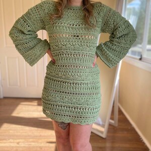 May include: A sage green crochet mini dress with long sleeves and a loose fit. The dress features a delicate openwork pattern and a scalloped hem.