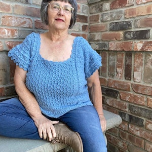 May include: A woman wearing a light blue crocheted top with a scalloped neckline and short sleeves. She is sitting on a stone bench with her legs crossed and wearing blue jeans and brown boots.