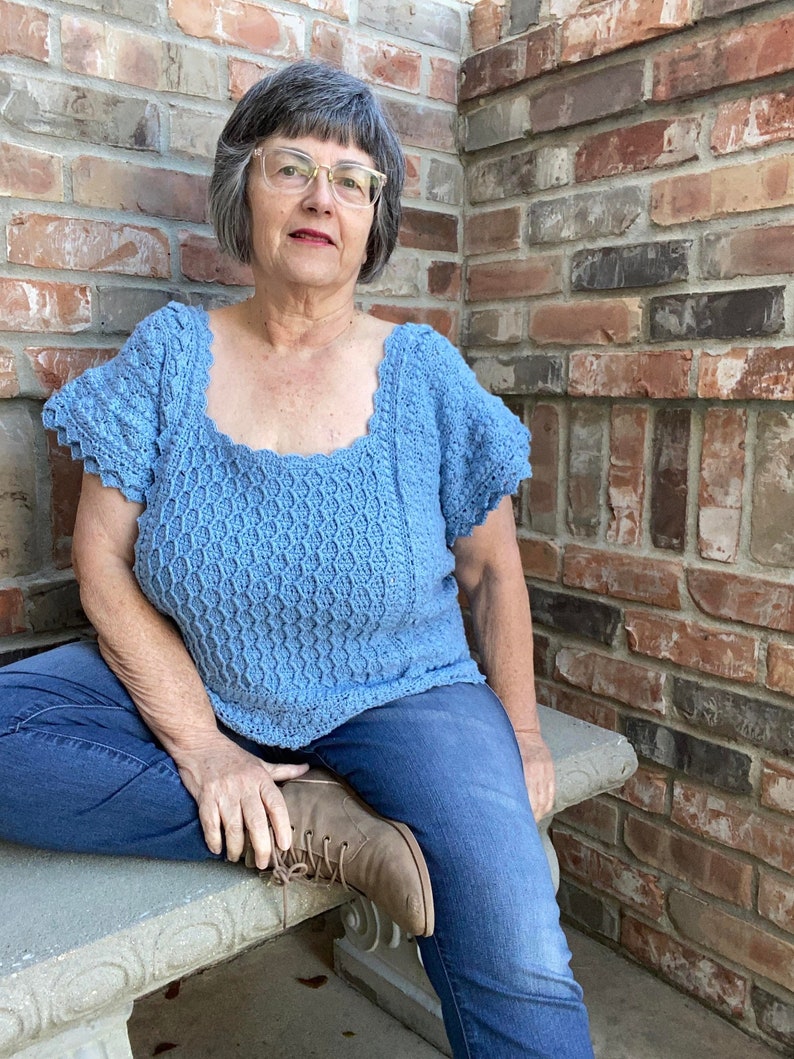 May include: A woman wearing a light blue crocheted top with a scalloped neckline and short sleeves. She is sitting on a stone bench with her legs crossed and wearing blue jeans and brown boots.