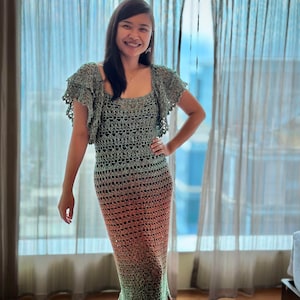 May include: A woman wearing a long, green, orange, and brown crochet dress with a ruffled neckline. The dress has a loose fit and is made of a lightweight, breathable fabric.