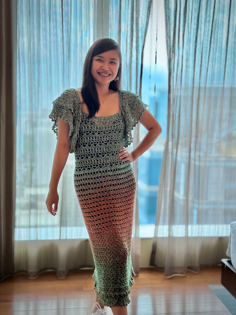 May include: A woman wearing a long, green, orange, and brown crochet dress with a ruffled neckline. The dress has a loose fit and is made of a lightweight, breathable fabric.