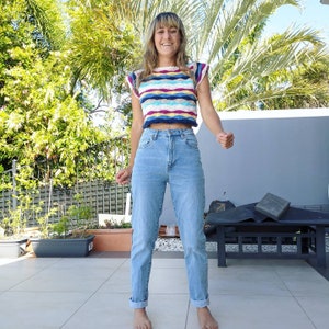 Op de afbeelding: Een vrouw draagt een kleurrijk gestreept gehaakt topje en lichtblauwe jeans. Het topje heeft een golvend patroon met tinten blauw, roze, wit en geel.