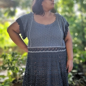 May include: A woman wearing a gray crochet dress with a lace pattern. The dress has a fitted bodice and a flowing skirt. The woman is standing in front of a blurred background of trees.