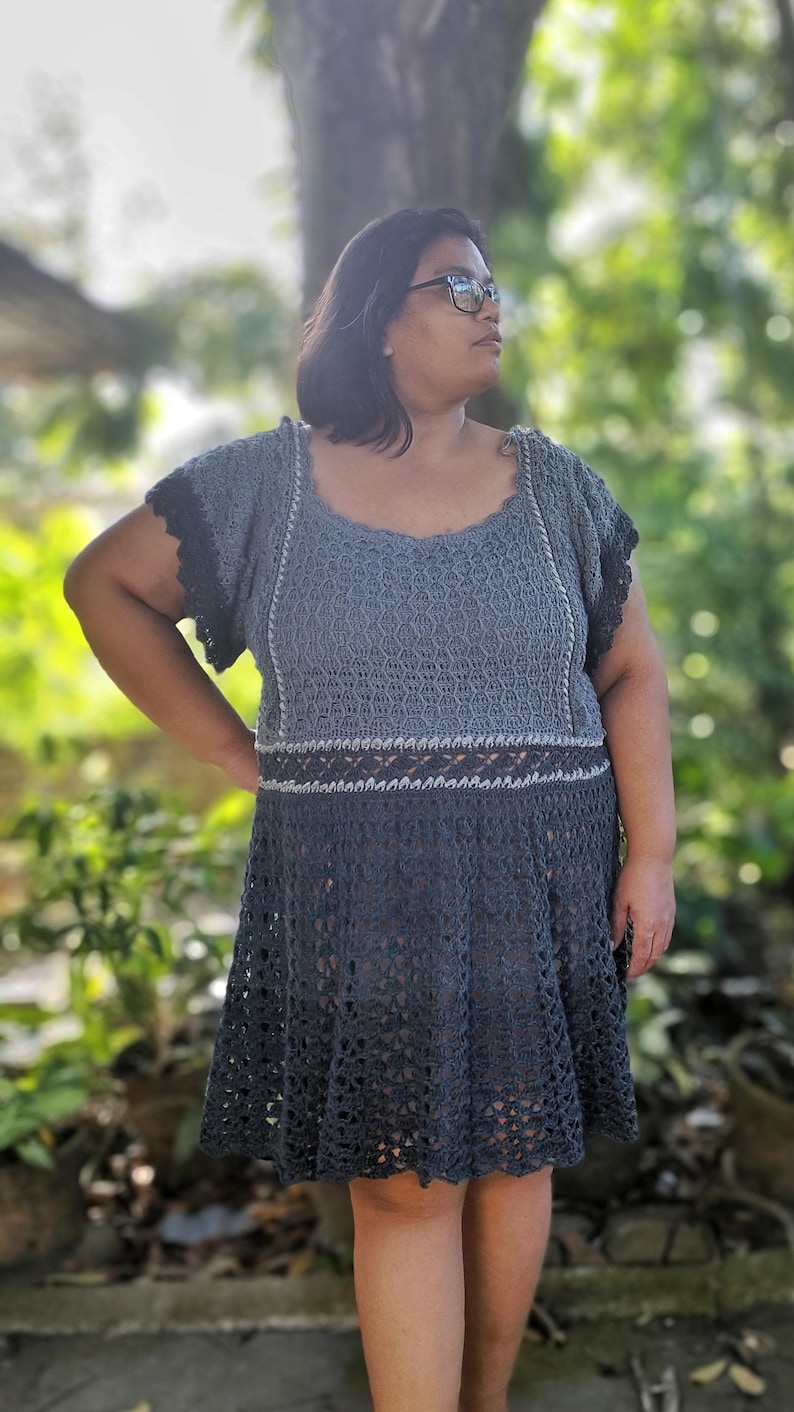 May include: A woman wearing a gray crochet dress with a lace pattern. The dress has a fitted bodice and a flowing skirt. The woman is standing in front of a blurred background of trees.