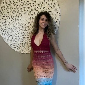 May include: A woman wearing a long, crocheted dress with a rainbow ombre pattern. The dress has a halter neckline and a fitted silhouette. The dress is made of a soft, lightweight yarn and is perfect for warm weather.