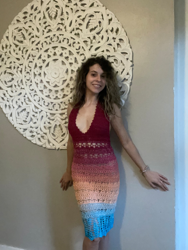 May include: A woman wearing a long, crocheted dress with a rainbow ombre pattern. The dress has a halter neckline and a fitted silhouette. The dress is made of a soft, lightweight yarn and is perfect for warm weather.