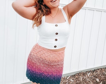 May include: A woman wearing a white crop top with buttons and a colorful crochet skirt. The skirt is a gradient of pink, purple, and blue.