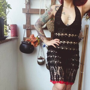 May include: A woman wearing a black crochet halter dress with red trim. She has red hair and tattoos on her arms. The dress is a crochet design with a lot of open space. The woman is standing on a balcony with plants and a wooden lattice wall behind her.