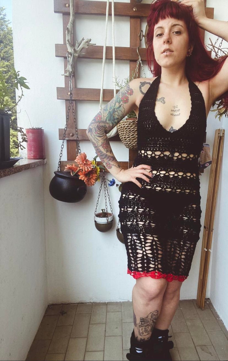 May include: A woman wearing a black crochet halter dress with red trim. She has red hair and tattoos on her arms. The dress is a crochet design with a lot of open space. The woman is standing on a balcony with plants and a wooden lattice wall behind her.