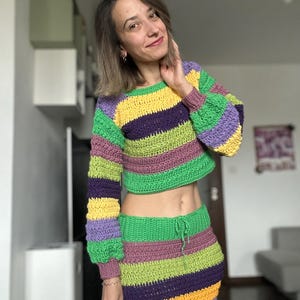 K&ouml;nnte beinhalten: Ein zweiteiliges H&auml;kelset mit einem gecroppten Langarmpullover und einem Minirock. Das Set zeigt horizontale Streifen in Gr&uuml;n, Lila, Gelb und Rosa. Der Pullover hat einen Rundhalsausschnitt und der Rock hat einen Kordelzug in der Taille.