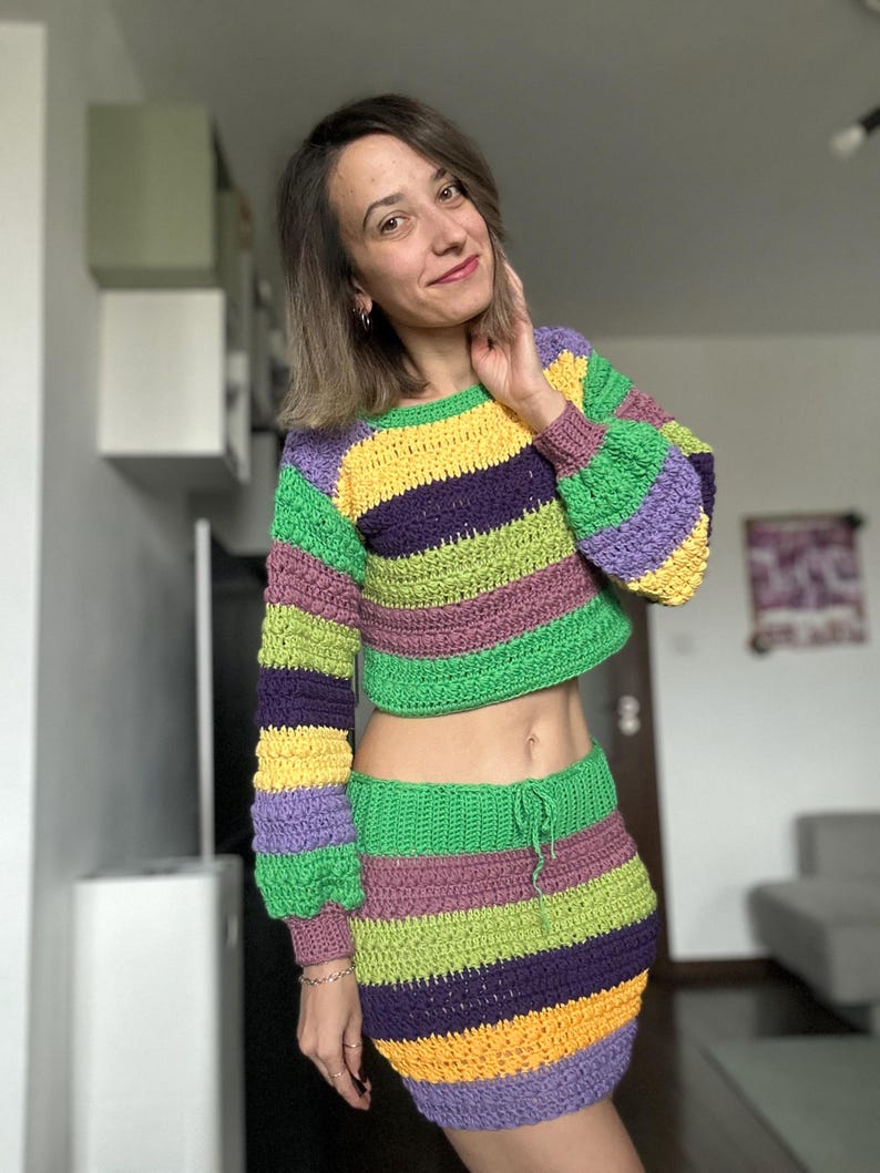 K&ouml;nnte beinhalten: Ein zweiteiliges H&auml;kelset mit einem gecroppten Langarmpullover und einem Minirock. Das Set zeigt horizontale Streifen in Gr&uuml;n, Lila, Gelb und Rosa. Der Pullover hat einen Rundhalsausschnitt und der Rock hat einen Kordelzug in der Taille.