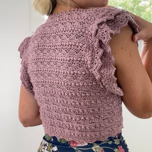 May include: A mauve crocheted top with a scalloped hem and ruffled sleeves. The top is sleeveless and has a close-fitting style.