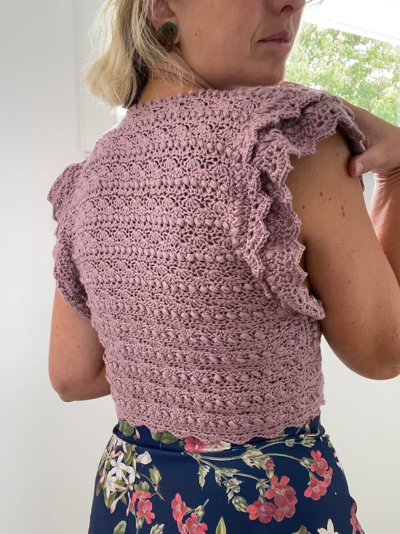 May include: A mauve crocheted top with a scalloped hem and ruffled sleeves. The top is sleeveless and has a close-fitting style.