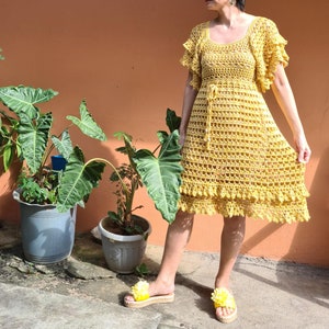 May include: A yellow crochet dress with a ruffled hem and short sleeves. The dress has a crocheted belt and is worn by a person standing on a patio.