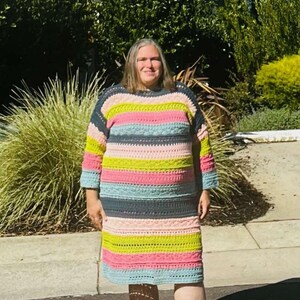 May include: A woman wearing a colourful striped crochet dress with a light blue, pink, green and dark grey colour scheme. She is standing on a paved surface in front of a house.