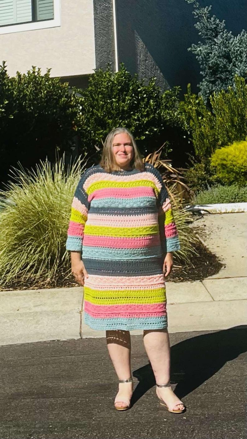 May include: A woman wearing a colourful striped crochet dress with a light blue, pink, green and dark grey colour scheme. She is standing on a paved surface in front of a house.