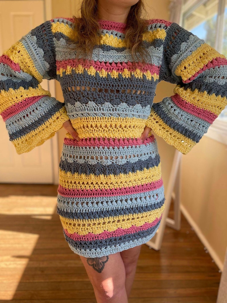 May include: A colourful crochet dress with a striped pattern in shades of yellow, pink, blue, and grey. The dress has a loose fit and long sleeves with a bell shape.