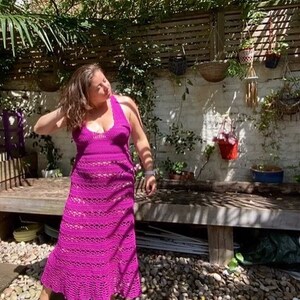 May include: A woman wearing a long, magenta crochet dress with a halter neckline. She is standing in a garden setting with a wooden bench and gravel.