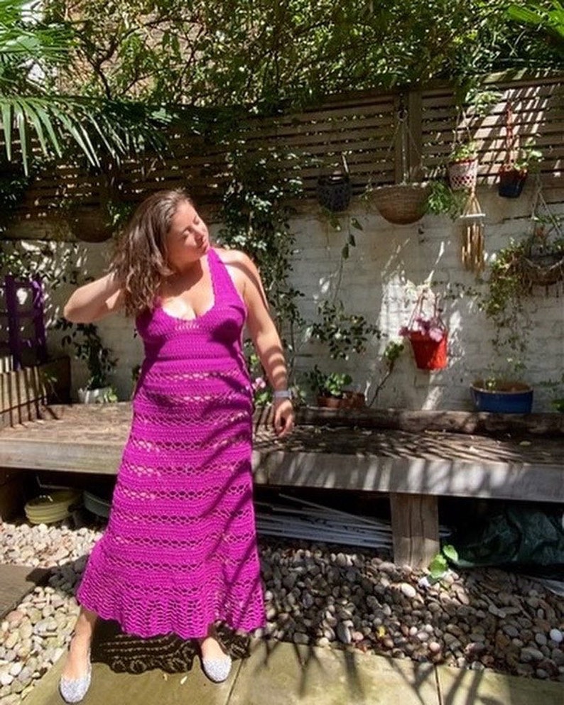 May include: A woman wearing a long, magenta crochet dress with a halter neckline. She is standing in a garden setting with a wooden bench and gravel.