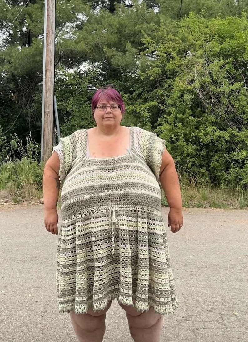 May include: A woman wearing a crocheted dress with a green and white striped pattern. The dress has a scoop neck and short sleeves. The woman has pink hair and is wearing glasses.