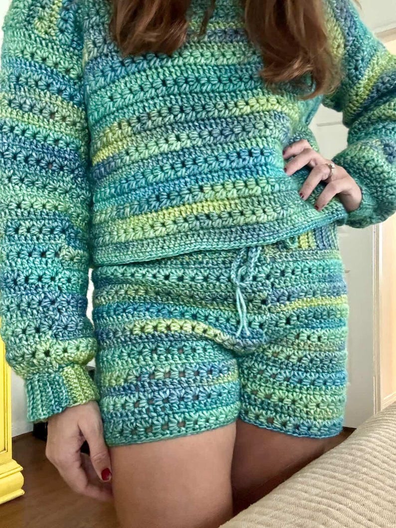 K&ouml;nnte beinhalten: Handgefertigtes H&auml;kelset in T&uuml;rkis-, Blau- und Gr&uuml;nt&ouml;nen. Das Set besteht aus einem lang&auml;rmeligen Pullover und passenden Shorts mit Kordelzug. Das H&auml;kelmuster weist ein strukturiertes Stichdesign auf.