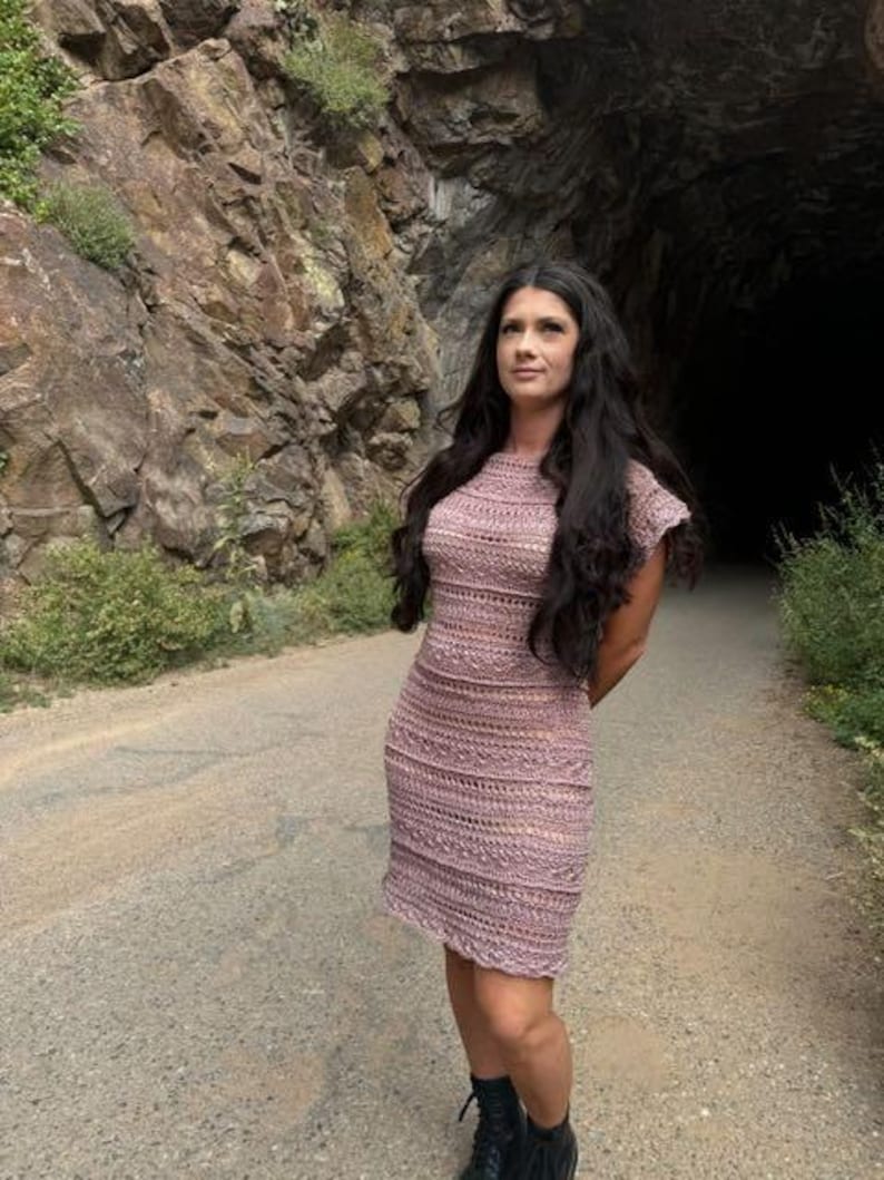 May include: A woman wearing a light pink crochet dress with a delicate pattern. She is standing in front of a rocky tunnel entrance.