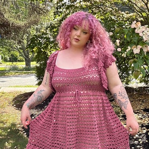 May include: A woman wearing a pink crocheted dress with a square neckline and a tie at the waist. The dress has a scalloped hem and is made with a delicate open stitch pattern. The woman has pink curly hair and is standing in front of a green bush with white flowers.
