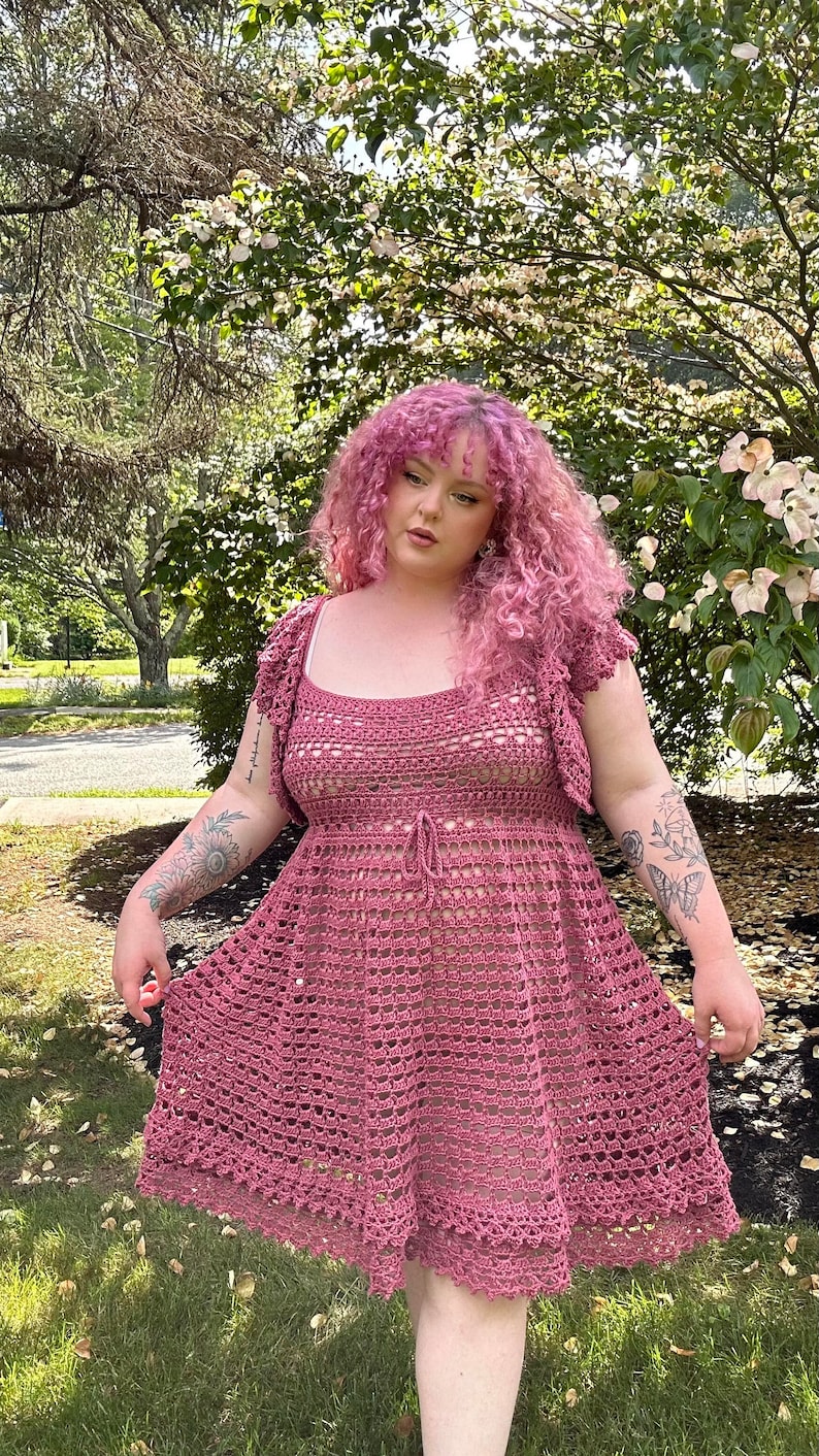 May include: A woman wearing a pink crocheted dress with a square neckline and a tie at the waist. The dress has a scalloped hem and is made with a delicate open stitch pattern. The woman has pink curly hair and is standing in front of a green bush with white flowers.
