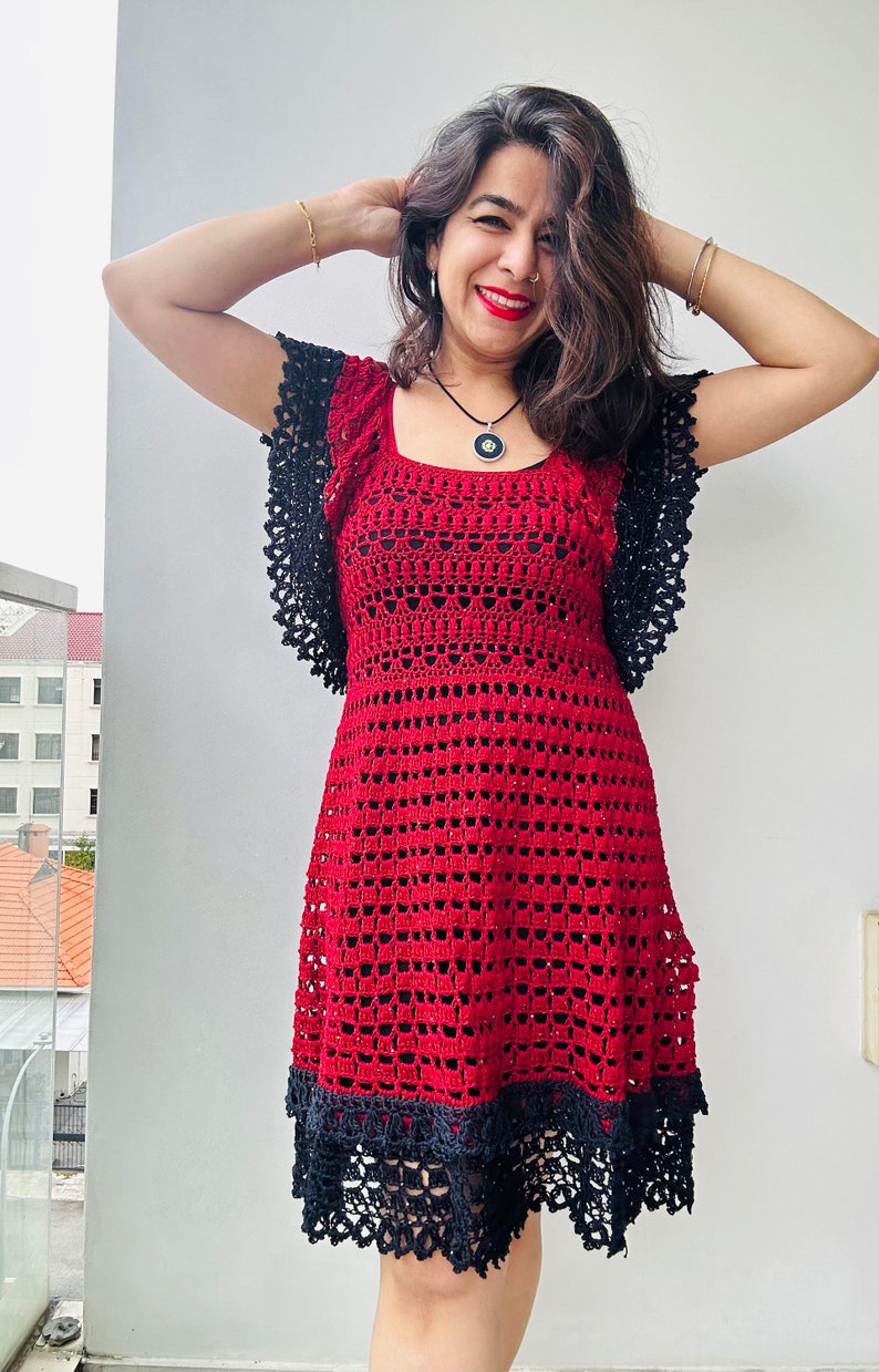 May include: A red crochet dress with black lace trim and short sleeves. The dress has a square neckline and a fitted bodice. The skirt is flared and falls to the knees.