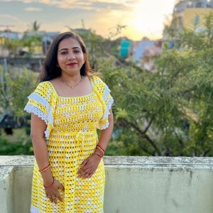 May include: A woman wearing a yellow and white crochet dress with a white crocheted trim. The dress has a square neckline and short sleeves.