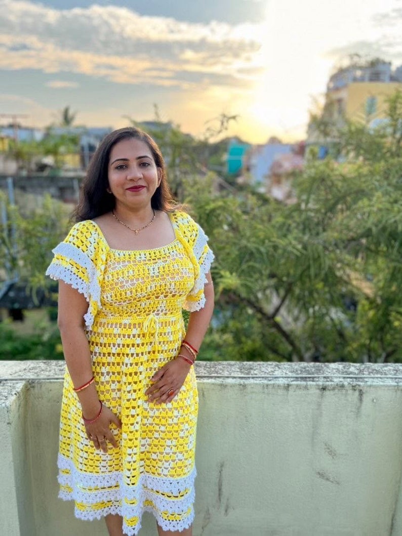 May include: A woman wearing a yellow and white crochet dress with a white crocheted trim. The dress has a square neckline and short sleeves.