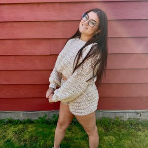 K&ouml;nnte beinhalten: Beige H&auml;kelpullover und Shorts Set. Der lang&auml;rmlige Pullover ist cropped, und die Shorts sind hoch tailliert. Das Set ist aus einem grob gestrickten Material gefertigt. Der Hintergrund ist eine rote Holzwand und gr&uuml;nes Gras.