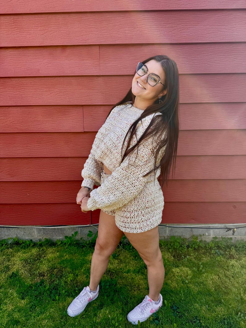 K&ouml;nnte beinhalten: Beige H&auml;kelpullover und Shorts Set. Der lang&auml;rmlige Pullover ist cropped, und die Shorts sind hoch tailliert. Das Set ist aus einem grob gestrickten Material gefertigt. Der Hintergrund ist eine rote Holzwand und gr&uuml;nes Gras.