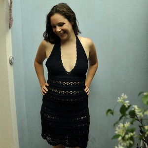 May include: A woman wearing a black crochet dress with a halter neckline. The dress has a fitted bodice and a flowing skirt.