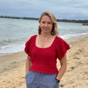 May include: A woman wearing a red crocheted top with a scalloped neckline and short sleeves, paired with blue linen pants. She is standing on a sandy beach with the ocean in the background.