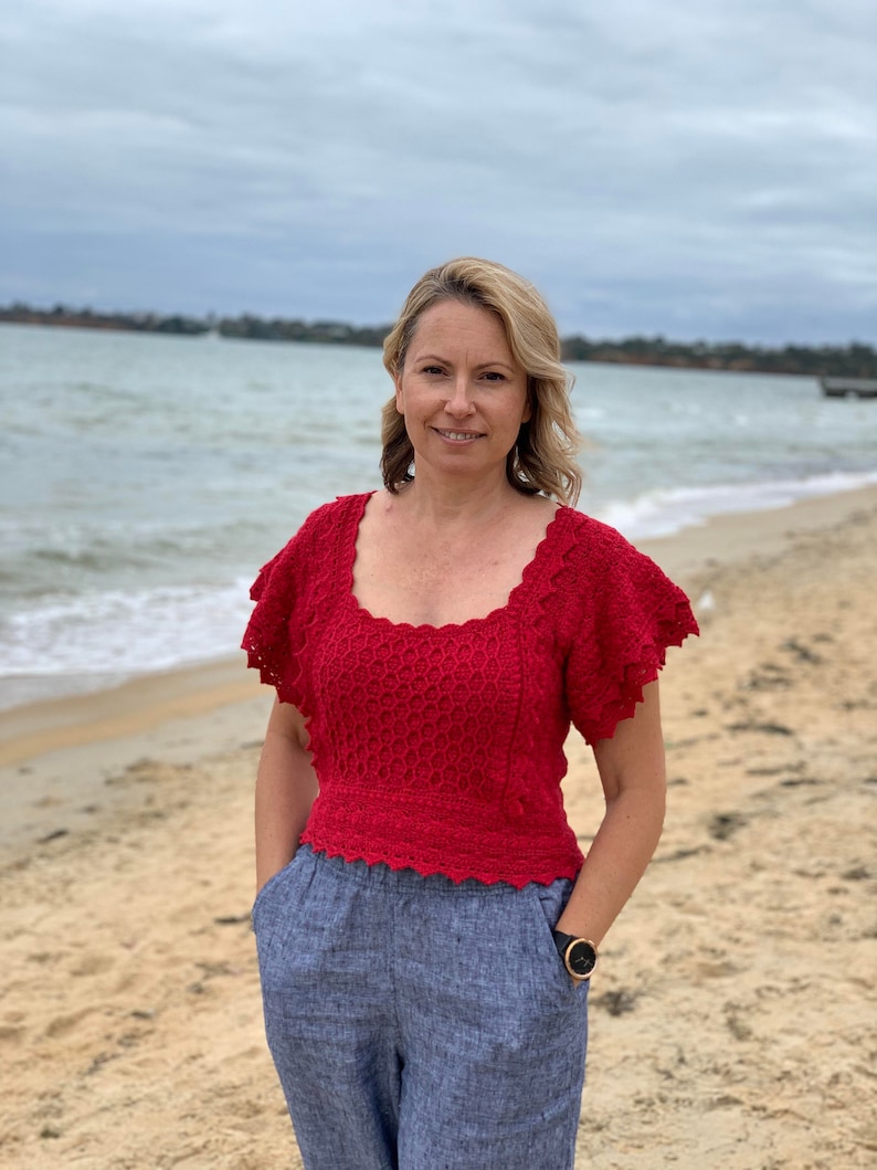 May include: A woman wearing a red crocheted top with a scalloped neckline and short sleeves, paired with blue linen pants. She is standing on a sandy beach with the ocean in the background.