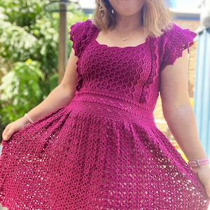 May include: A magenta crochet dress with a square neckline and short, ruffled sleeves. The dress has a fitted bodice and a flowing skirt.