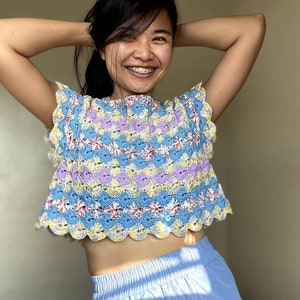 May include: A woman wearing a colorful crocheted crop top with a scalloped hem. The top features a blue, yellow, purple, and red color scheme.