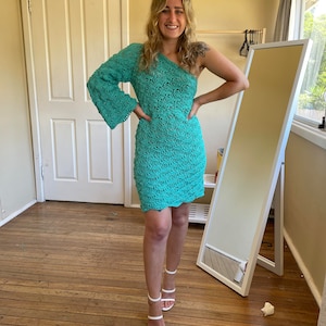 May include: A woman wearing a teal crochet dress with one long sleeve and a short sleeve on the other side. She is wearing white heeled sandals and is standing on a wooden floor.