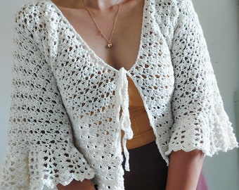 May include: White crochet cardigan with a tie front and bell sleeves. The cardigan is made of a delicate, open-stitch pattern.