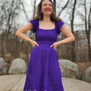May include: A woman wearing a purple crochet dress with a square neckline and short sleeves. The dress has a fitted bodice and a flowing skirt.
