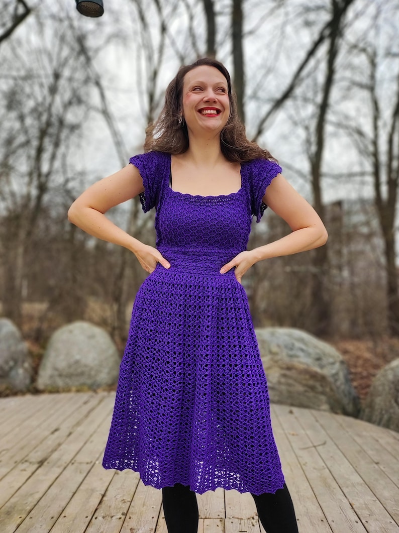 May include: A woman wearing a purple crochet dress with a square neckline and short sleeves. The dress has a fitted bodice and a flowing skirt.