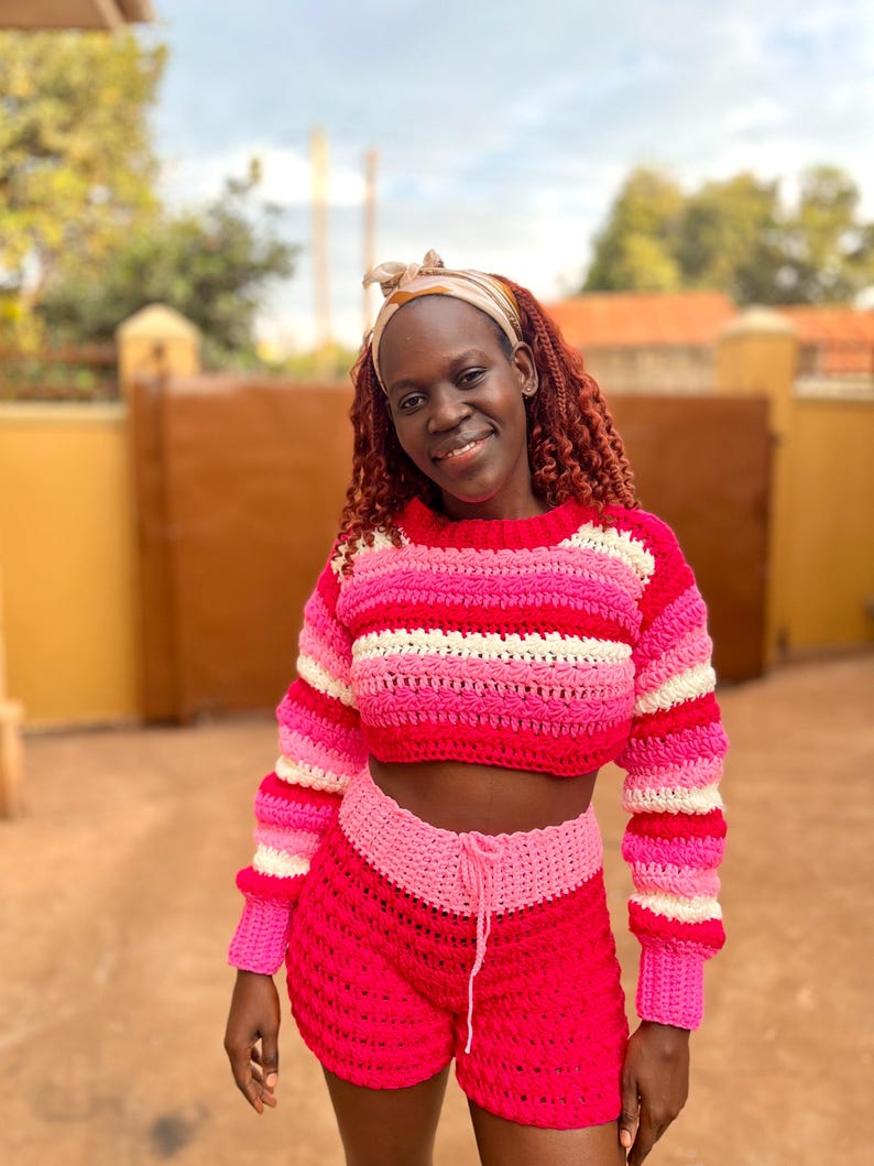 K&ouml;nnte beinhalten: Handgefertigtes H&auml;kel-Crop-Top und Shorts-Set in Rosa- und Wei&szlig;t&ouml;nen. Das Oberteil hat horizontale Streifen, w&auml;hrend die Shorts einen Kordelzug in der Taille haben. Ein farbenfrohes Beispiel f&uuml;r H&auml;kelbekleidung.