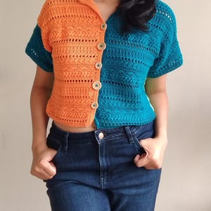 May include: A crocheted top with a button-up front, featuring a split colour design with orange and teal. The top has short sleeves and a cropped length.